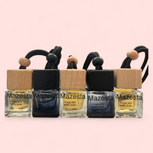 Puede incluir: Cinco pequeños frascos de vidrio con tapas de madera y tapas negras. Cada frasco tiene una cuerda negra atada a la tapa y contiene un líquido transparente. Los frascos están etiquetados con "Mazesta" y "From MD with love".