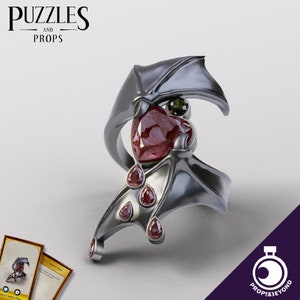 May include: A silver ring with a bat wing design and red and green gemstones. The ring is a statement piece with a gothic aesthetic.
