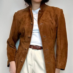 May include: A brown suede jacket with a mandarin collar and button closure. The jacket has a long, fitted silhouette and is worn over a white t-shirt and cream-colored pants.
