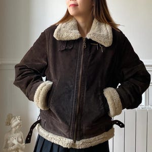 May include: A brown suede jacket with a cream-colored faux fur collar and trim. The jacket features a zip-up front, two pockets, and adjustable straps at the waist. The jacket is worn over a black pleated skirt.