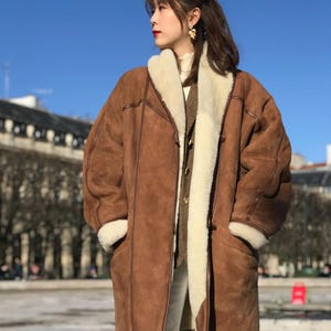 May include: A brown shearling coat with a cream-colored shearling lining and trim. The coat has a long length, large pockets, and a button closure. The person is wearing white pants and a cream-colored sweater.