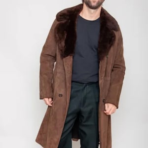 May include: A long brown suede coat with a dark brown fur collar. The coat has a double-breasted design and is worn with black pants and shoes. The coat is a vintage style.