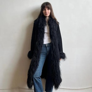 May include: A long black coat with a shaggy fur trim along the collar, front, and cuffs. The coat is worn over a white top and blue jeans. The model is wearing black boots. The background is a plain white wall and wooden floor.