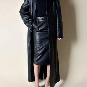 May include: A full-length black leather coat, paired with a matching leather skirt and top. The coat has a classic design with a lapel collar and a belt. The outfit is completed with beige heels.