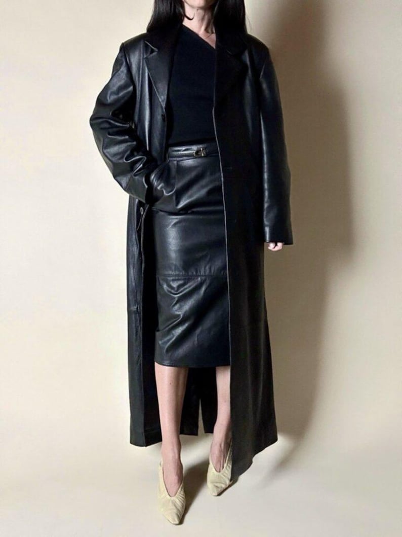 May include: A full-length black leather coat, paired with a matching leather skirt and top. The coat has a classic design with a lapel collar and a belt. The outfit is completed with beige heels.