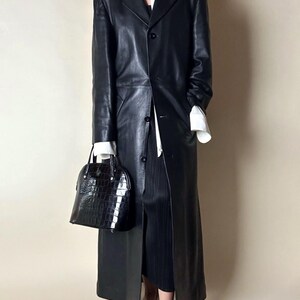 May include: A long, black leather coat with a classic design. The coat features a button-down front and a tailored fit. A black, textured handbag is held in the hand. The outfit is completed with a white shirt and black skirt.