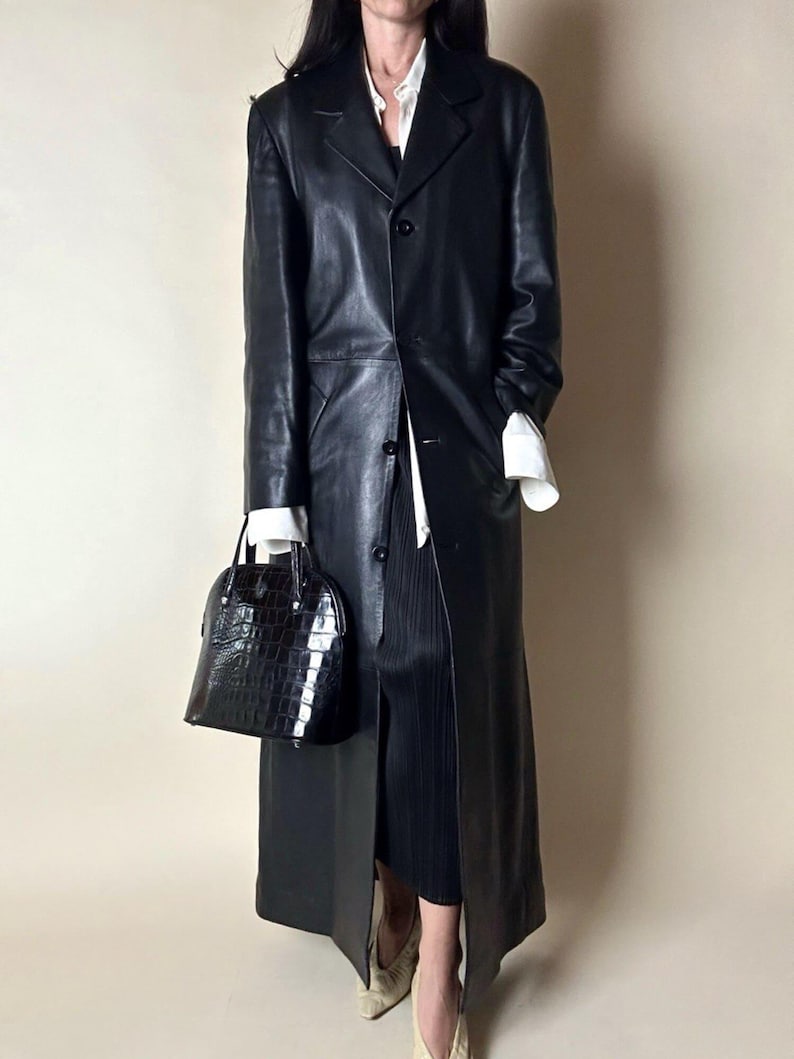 May include: A long, black leather coat with a classic design. The coat features a button-down front and a tailored fit. A black, textured handbag is held in the hand. The outfit is completed with a white shirt and black skirt.