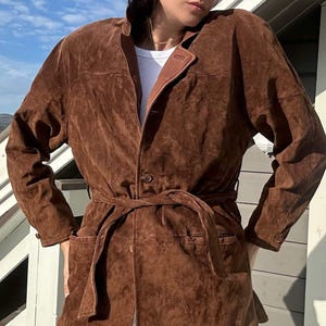 May include: A brown suede jacket with a belt and a button fastening. The jacket has a slightly oversized fit and a relaxed silhouette.