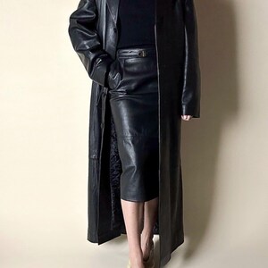 May include: A full-length black leather coat with a matching leather skirt. The coat has a classic trench coat design with a button closure. The skirt is knee-length and has a belt. The outfit is completed with beige shoes.