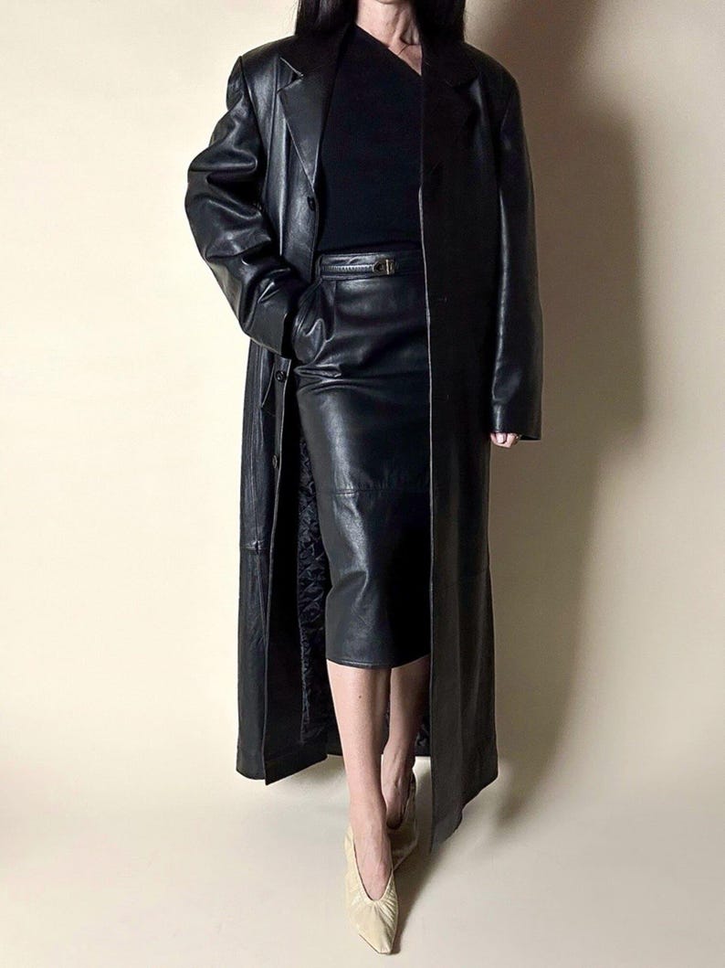 May include: A full-length black leather coat with a matching leather skirt. The coat has a classic trench coat design with a button closure. The skirt is knee-length and has a belt. The outfit is completed with beige shoes.