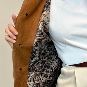 May include: A brown suede coat with a patterned lining. The lining is a mix of brown, white, blue, and red colors. The coat has a button closure.
