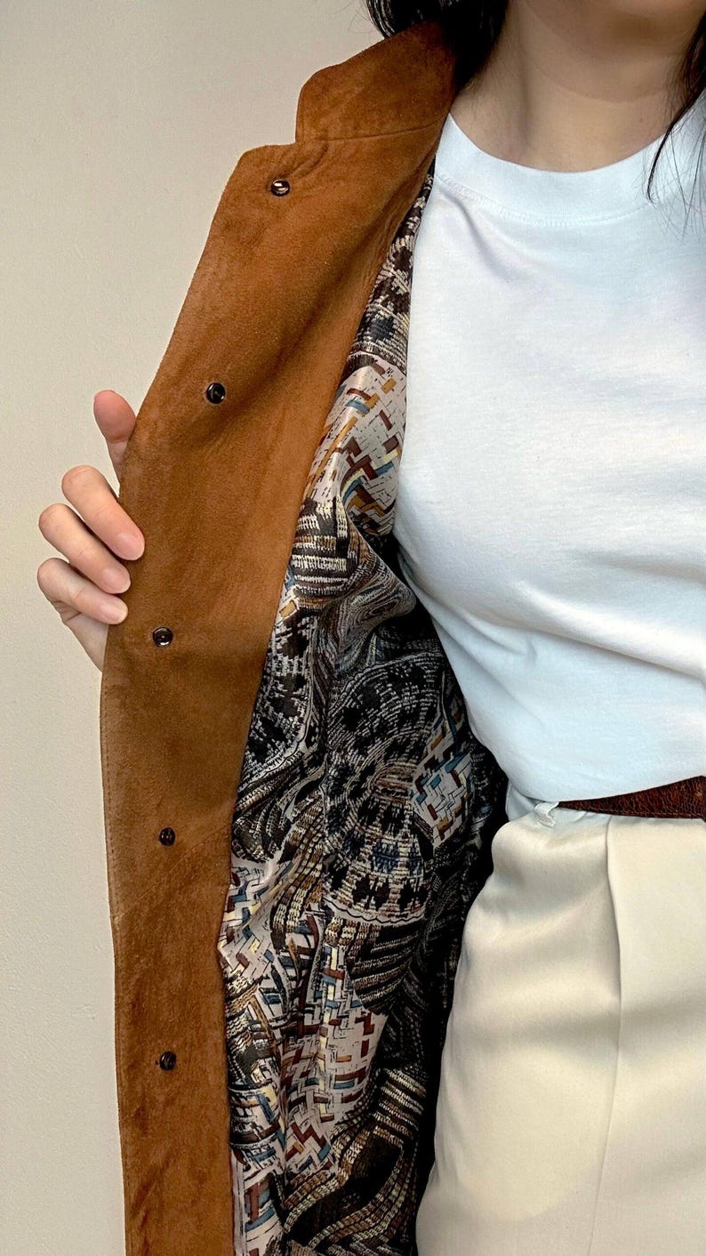 May include: A brown suede coat with a patterned lining. The lining is a mix of brown, white, blue, and red colors. The coat has a button closure.
