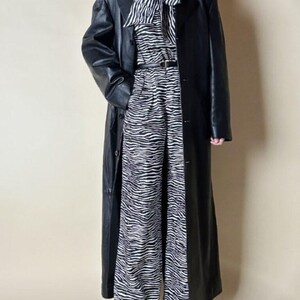 May include: A long black leather coat over a zebra-print outfit. The outfit includes a zebra-striped blouse with a bow, matching wide-leg pants, and a thin black belt. The coat is buttoned and reaches the ankles.