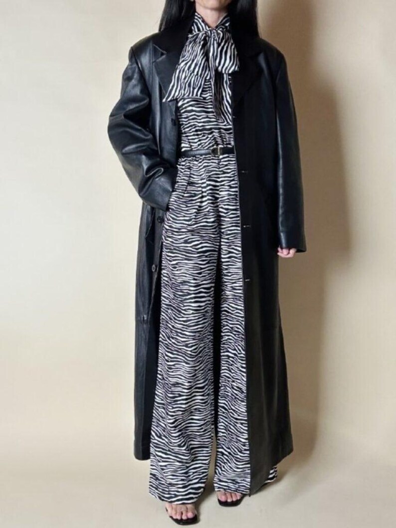 May include: A long black leather coat over a zebra-print outfit. The outfit includes a zebra-striped blouse with a bow, matching wide-leg pants, and a thin black belt. The coat is buttoned and reaches the ankles.
