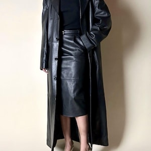 May include: A long, black leather coat with a matching skirt. The coat is buttoned and reaches the ankles. The skirt is knee-length. The outfit is paired with beige heels. The overall style is classic and elegant.
