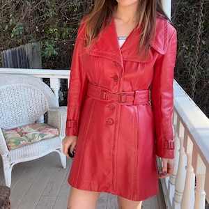 Puede incluir: Una gabardina de cuero roja con cinturón y cierre de botones. La gabardina tiene cuello y mangas largas.