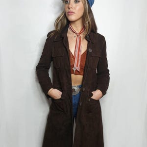 Op de afbeelding: Een lange bruine su&egrave;de jas met knoopsluiting en zakken. De jas wordt gedragen over een rood-wit gestreepte sjaal en een blauwe denim pet.