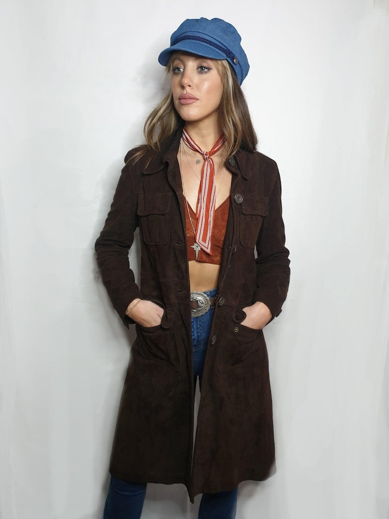 Op de afbeelding: Een lange bruine su&egrave;de jas met knoopsluiting en zakken. De jas wordt gedragen over een rood-wit gestreepte sjaal en een blauwe denim pet.