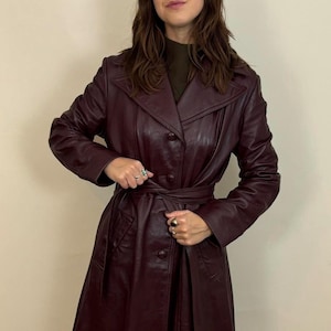 Peut inclure: Un trench-coat en cuir bordeaux avec une taille ceinturée et des boutons. Le manteau a un col à revers classique et des manches longues. La couleur du manteau est profonde et riche.