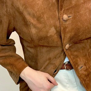 May include: A brown suede jacket with a mandarin collar and button closure. The jacket has a fitted silhouette and long sleeves.