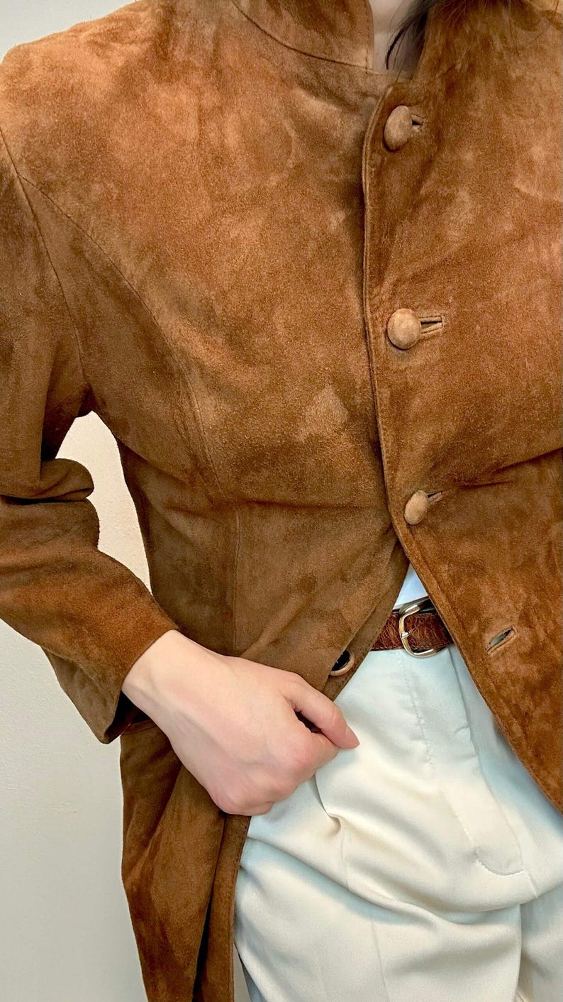 May include: A brown suede jacket with a mandarin collar and button closure. The jacket has a fitted silhouette and long sleeves.