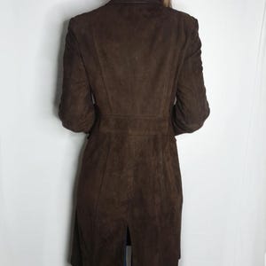 Op de afbeelding: Een lange bruine su&egrave;de jas met een split in de rug. De jas heeft een klassieke kraag en lange mouwen.
