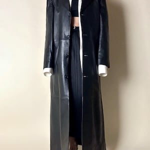 May include: A long, black leather coat with a classic lapel collar and button closure. The coat is floor-length and appears to be vintage. The model is wearing a white shirt and black top underneath.
