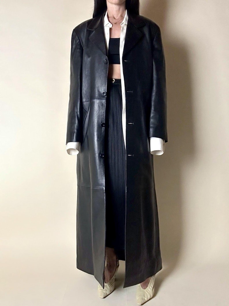May include: A long, black leather coat with a classic lapel collar and button closure. The coat is floor-length and appears to be vintage. The model is wearing a white shirt and black top underneath.