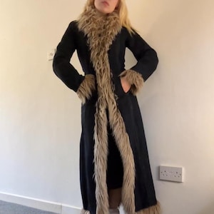 May include: A long black coat with faux fur trim and accents. The coat features a faux fur collar, cuffs, and trim down the front. The coat has pockets and is worn open. The coat is a vintage style.