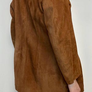 May include: A brown suede jacket with a long, relaxed fit. The jacket has a stand-up collar and long sleeves.
