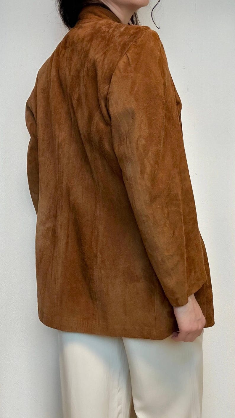May include: A brown suede jacket with a long, relaxed fit. The jacket has a stand-up collar and long sleeves.
