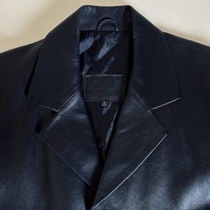 May include: A close-up of a black leather blazer. The blazer has a notched lapel, a single-button closure, and a small loop at the back of the collar. The lining is black. The blazer appears to be a size small.
