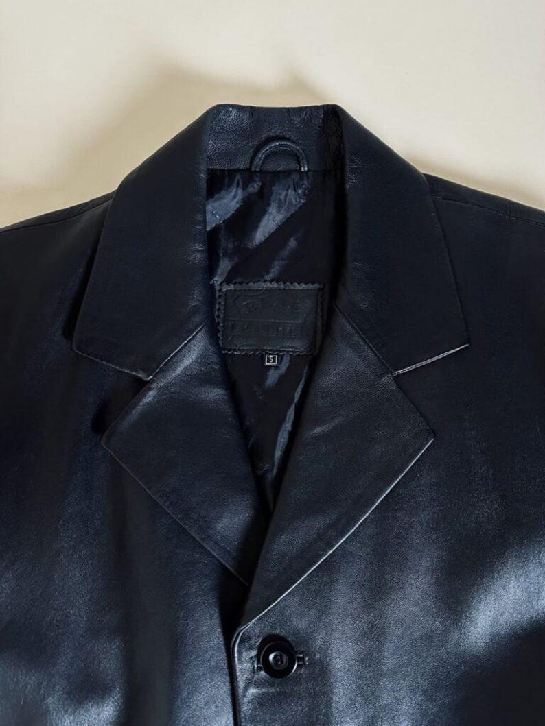 May include: A close-up of a black leather blazer. The blazer has a notched lapel, a single-button closure, and a small loop at the back of the collar. The lining is black. The blazer appears to be a size small.