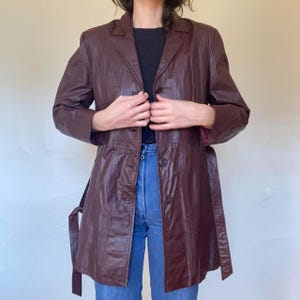 May include: A long, brown leather jacket with a belt and a button closure. The jacket has a collar and a slightly fitted silhouette.