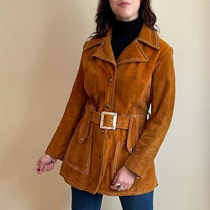 May include: A vintage brown suede jacket with a belted waist and large square buckle. The jacket features a collared neckline, button closures, and white stitching details. The jacket has two front pockets and is worn over a black turtleneck.