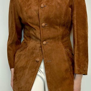 May include: A brown suede jacket with a mandarin collar and button closure. The jacket has a fitted silhouette and long sleeves.