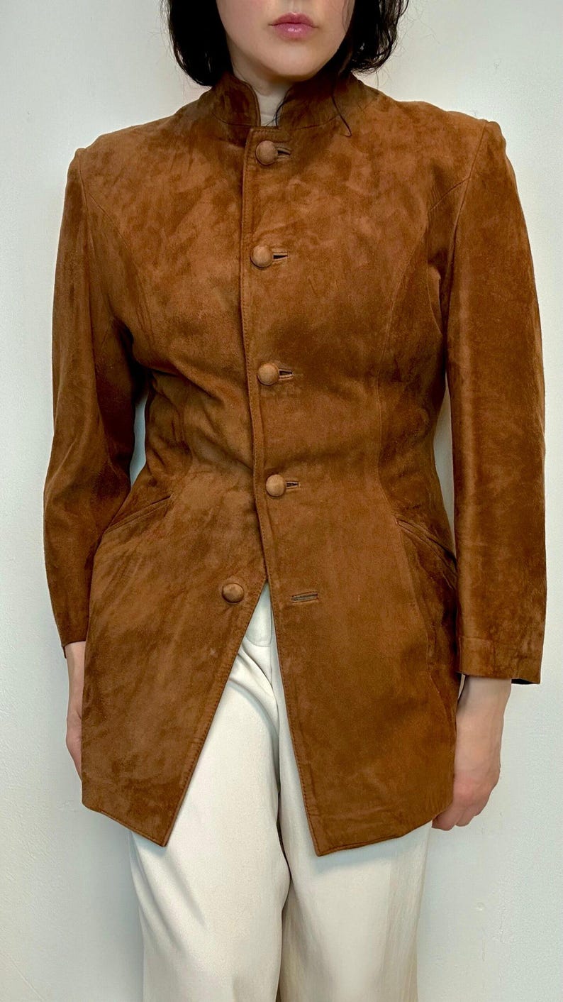 May include: A brown suede jacket with a mandarin collar and button closure. The jacket has a fitted silhouette and long sleeves.
