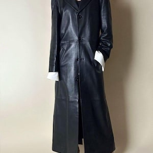 May include: A long, black leather coat with a classic design. The coat features a notched collar, button closure, and long sleeves. White cuffs peek out from the sleeves. The coat is floor-length and has a tailored fit, suitable for various occasions.