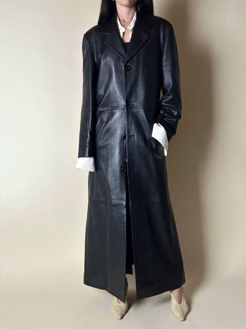 May include: A long, black leather coat with a classic design. The coat features a notched collar, button closure, and long sleeves. White cuffs peek out from the sleeves. The coat is floor-length and has a tailored fit, suitable for various occasions.