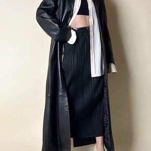 May include: A long black leather coat with a classic lapel collar. The coat is paired with a black pleated skirt, a white button-down shirt, and cream-colored heels. The outfit is completed with a black belt and a black top.