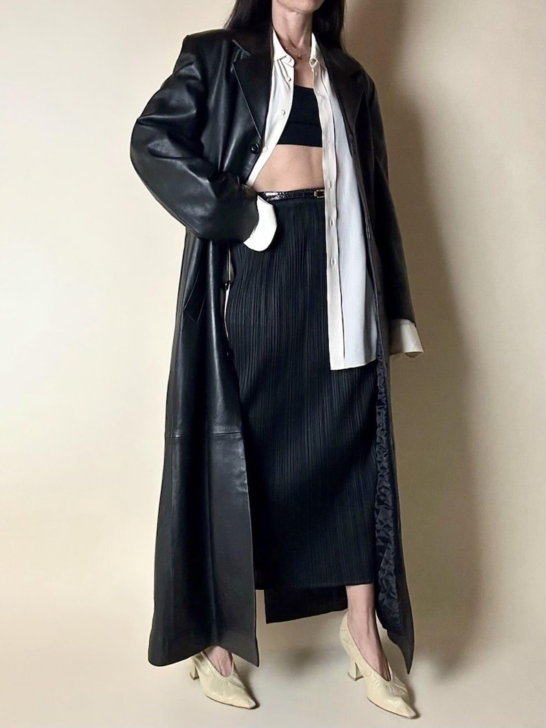 May include: A long black leather coat with a classic lapel collar. The coat is paired with a black pleated skirt, a white button-down shirt, and cream-colored heels. The outfit is completed with a black belt and a black top.