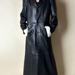 May include: A long, black leather coat with a button-up front and notched lapels. The coat has two front pockets and a slit in the back. The model is wearing a white shirt underneath.