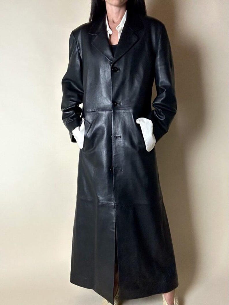 May include: A long, black leather coat with a button-up front and notched lapels. The coat has two front pockets and a slit in the back. The model is wearing a white shirt underneath.