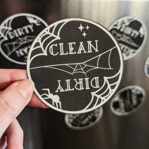 Puede incluir: Im&aacute;n redondo en blanco y negro con las palabras "CLEAN" y "DIRTY" en un dise&ntilde;o de telara&ntilde;a. Una peque&ntilde;a ara&ntilde;a est&aacute; en la parte inferior izquierda. El im&aacute;n est&aacute; sostenido por una mano.