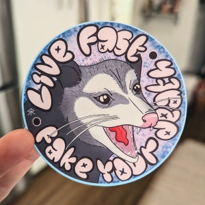 May include: A round sticker with a blue border and a pink background. The sticker features a cartoon opossum with a toothy grin and the text "Live Fast, Fake Your Death".