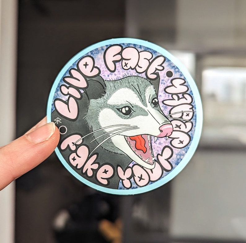 May include: A round sticker with a blue background and a white border. The sticker features a cartoon illustration of a possum with a pink nose and open mouth, with the text "Live Fast, Fake Your Death" in pink letters.