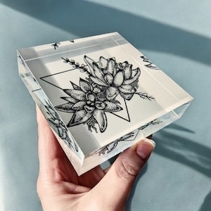 May include: A clear, square acrylic block featuring a black and white illustration of succulents and a geometric triangle. The artwork is detailed with stippling and fine lines, creating a botanical theme. The block is held against a light blue background.