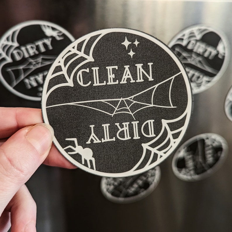 Puede incluir: Im&aacute;n redondo en blanco y negro con las palabras "CLEAN" y "DIRTY" en un dise&ntilde;o de telara&ntilde;a. Una peque&ntilde;a ara&ntilde;a est&aacute; en la parte inferior izquierda. El im&aacute;n est&aacute; sostenido por una mano.