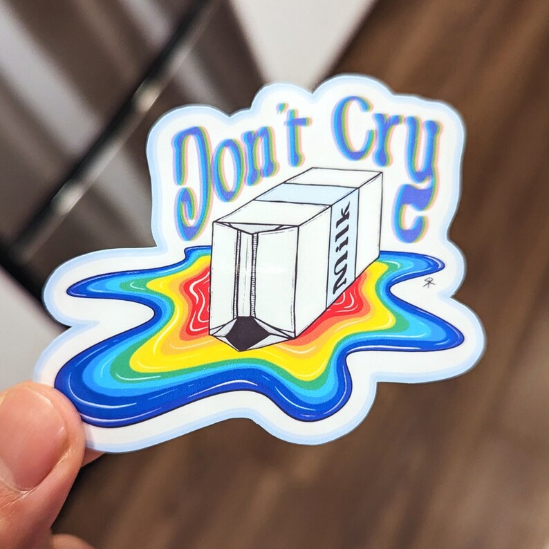 May include: A sticker with the words "Don't Cry" in a blue and purple font. The sticker features a carton of milk spilling a rainbow-colored liquid. The milk carton is white with the word "Milk" printed on the side.