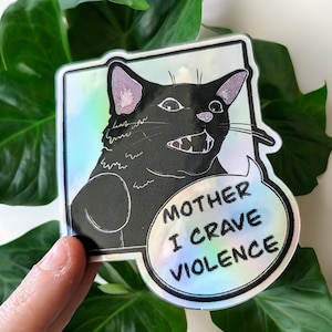 May include: A holographic sticker featuring a black cat with an angry expression and a speech bubble that says "Mother I crave violence".